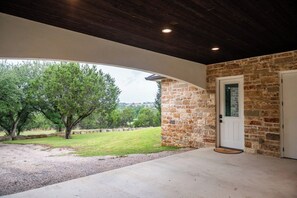A covered outdoor area with a stone wall and a door, a grassy lawn, and trees in the background. - Hill Country Reunion & Event Estate | Sleeps 28 (Fredericksburg)