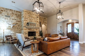 A living room with a stone wall, leather sofa, and a fireplace. - Hill Country Reunion & Event Estate | Sleeps 28 (Fredericksburg)