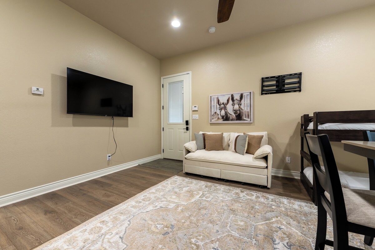 A room with a flat-screen TV mounted on the wall, a sofa with cushions, a dining table with chairs, and a patterned rug on the floor.