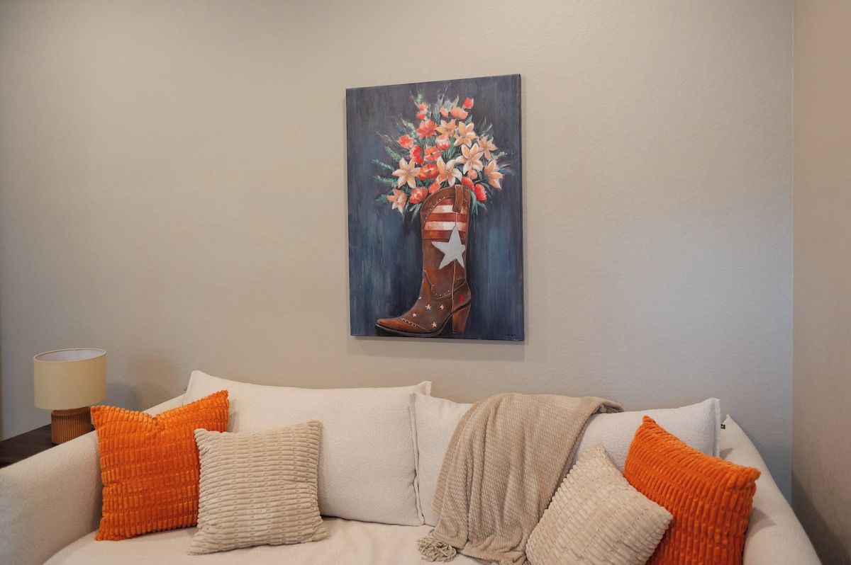 A living room with a couch, pillows, a lamp, and a painting of a boot with flowers.
