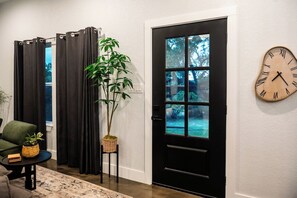 A room with a black door, a wall clock, a potted plant, a green chair, and a small table with a basket. - Modern Hilltop Farmhouse w/Patio in Wine Country (Fredericksburg)