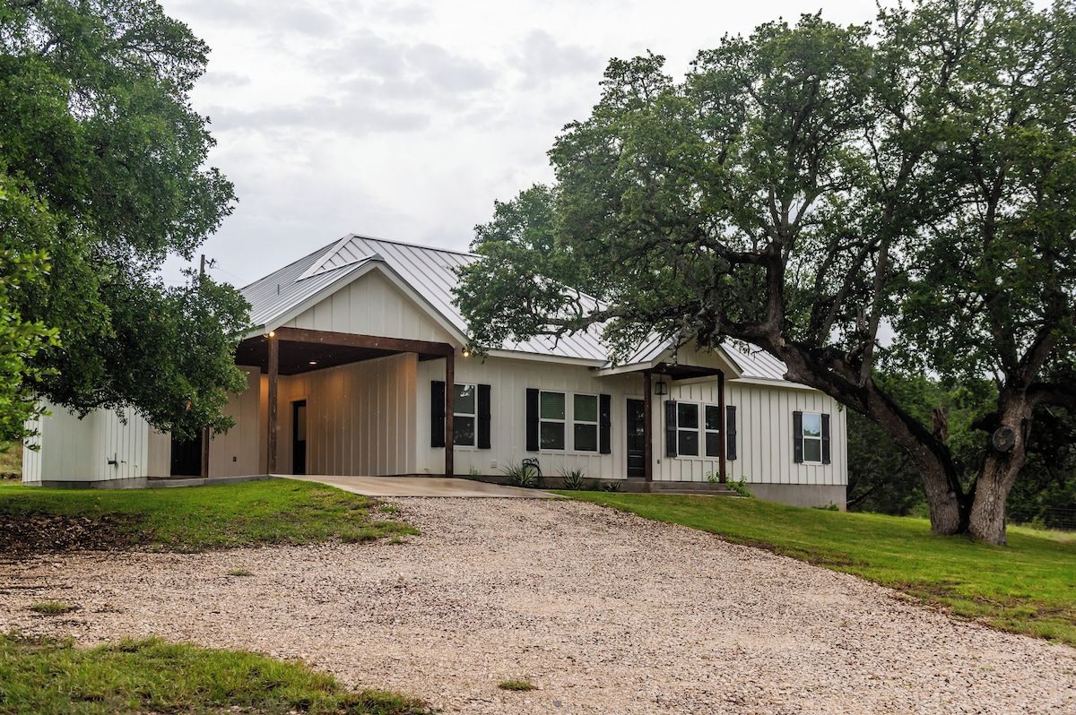 Una casa de una sola planta con porche cubierto, camino de grava y un árbol grande a la derecha.