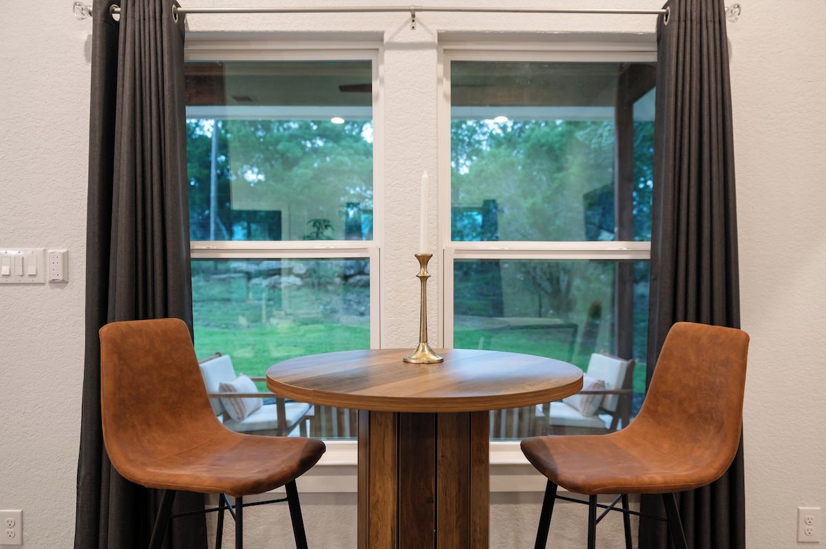 A round wooden table with two chairs, a candle, and a window with curtains.