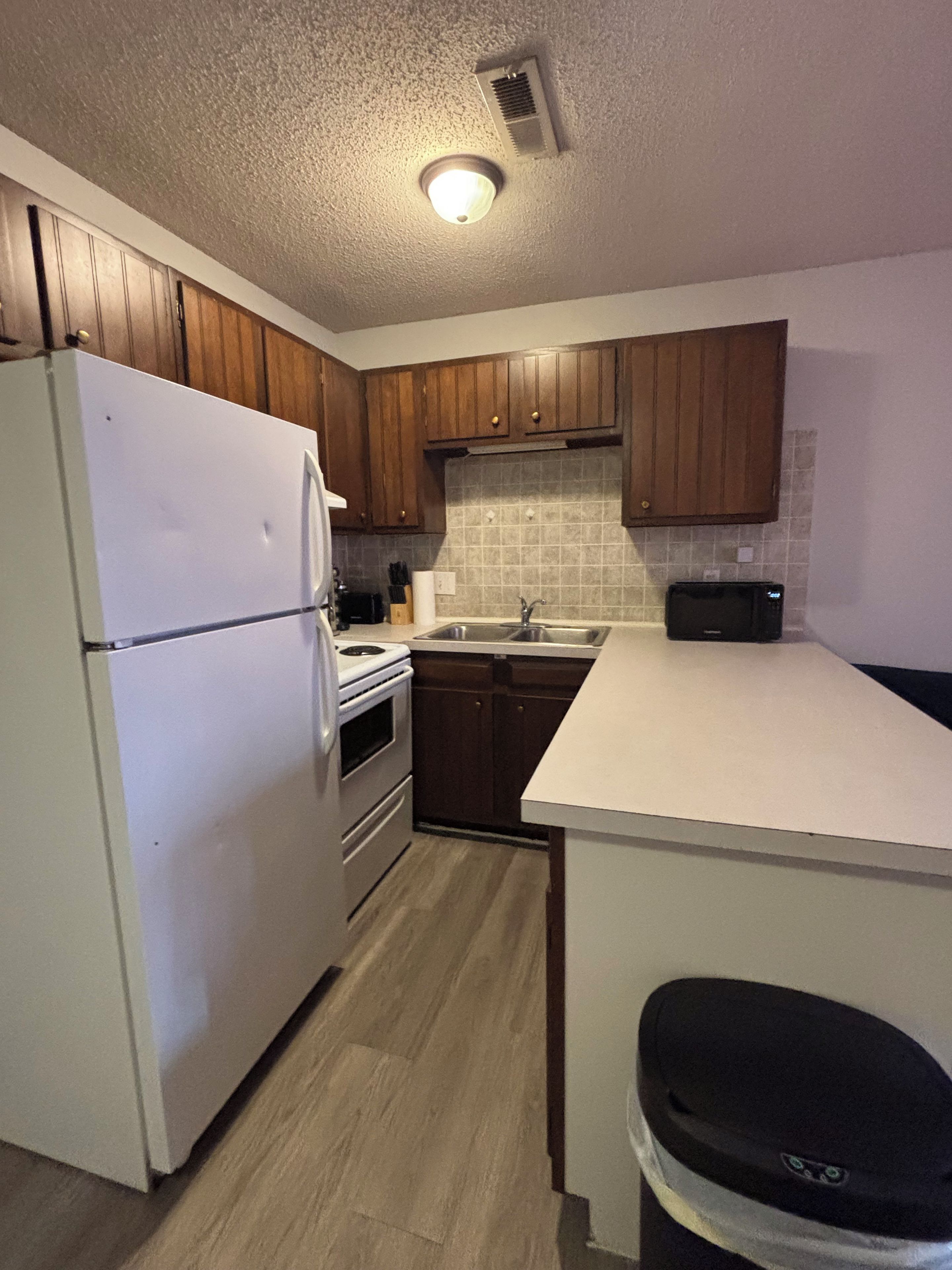 A kitchen with wooden cabinets, a white refrigerator, a stove, a sink, and a trash bin.