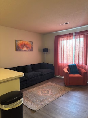 Un salon avec un canapé, un fauteuil, une lampe, un tableau et une fenêtre avec des rideaux.