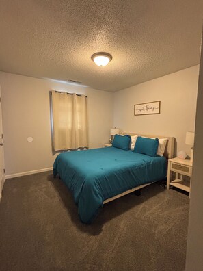 Une chambre à coucher comprenant un lit, une table de chevet, une lampe fixée au mur, une fenêtre avec des rideaux et un luminaire au plafond.