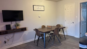 Een eethoek met een tafel, stoelen, een flatscreen-tv en een wandplank met een plant.