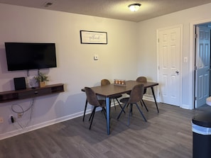 Un espace repas comprenant une table, des chaises, un téléviseur à écran plat et une étagère murale agrémentée d’une plante.
