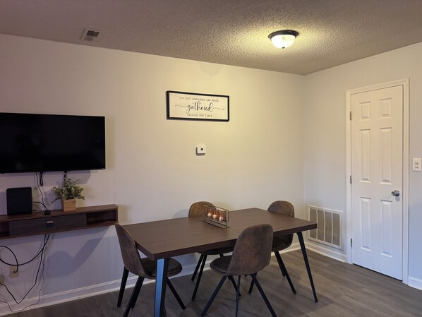 A dining area with a table, chairs, a flat-screen TV, a framed quote on the wall, and a door.