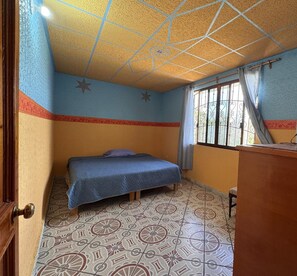 A small, single-bed room with patterned floor tiles, a wooden desk, and a window with metal bars.