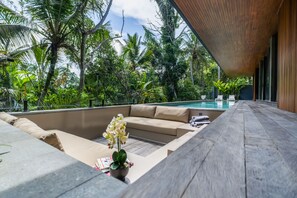 A poolside area with a wooden deck, beige sofa, and a small table with a potted plant. - Basic House (Gianyar)