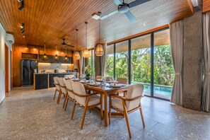 A modern dining area with a wooden table and chairs, a kitchen in the background, and a large window with a view of greenery. - Basic House (Gianyar)
