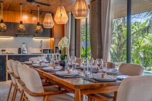 A dining area with a wooden table set for a meal, surrounded by chairs, with hanging pendant lights above. - Basic House (Gianyar)