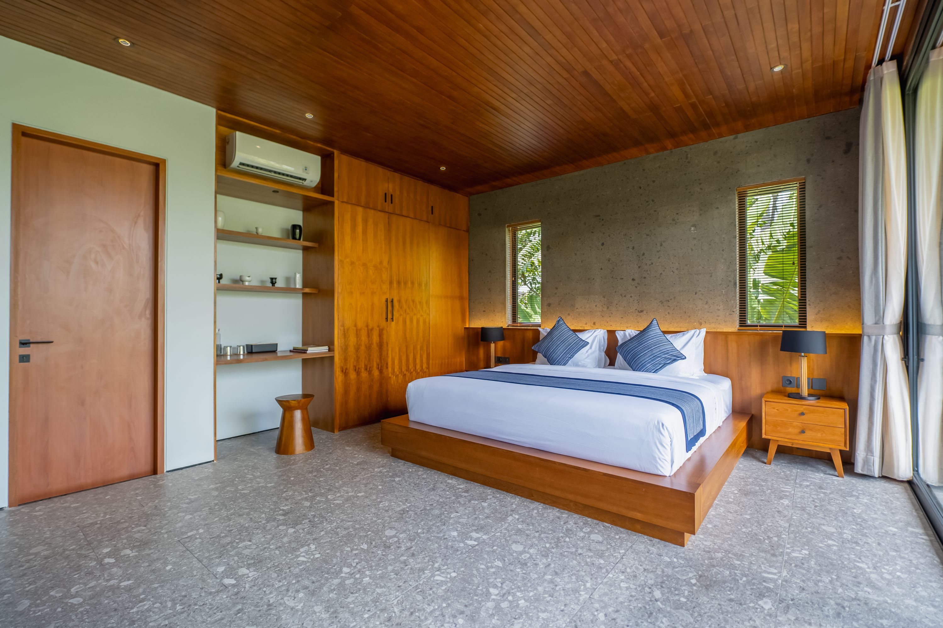 Een moderne slaapkamer met een houten plafond, een groot bed met blauw beddengoed, een inbouwkast en een klein nachtkastje.