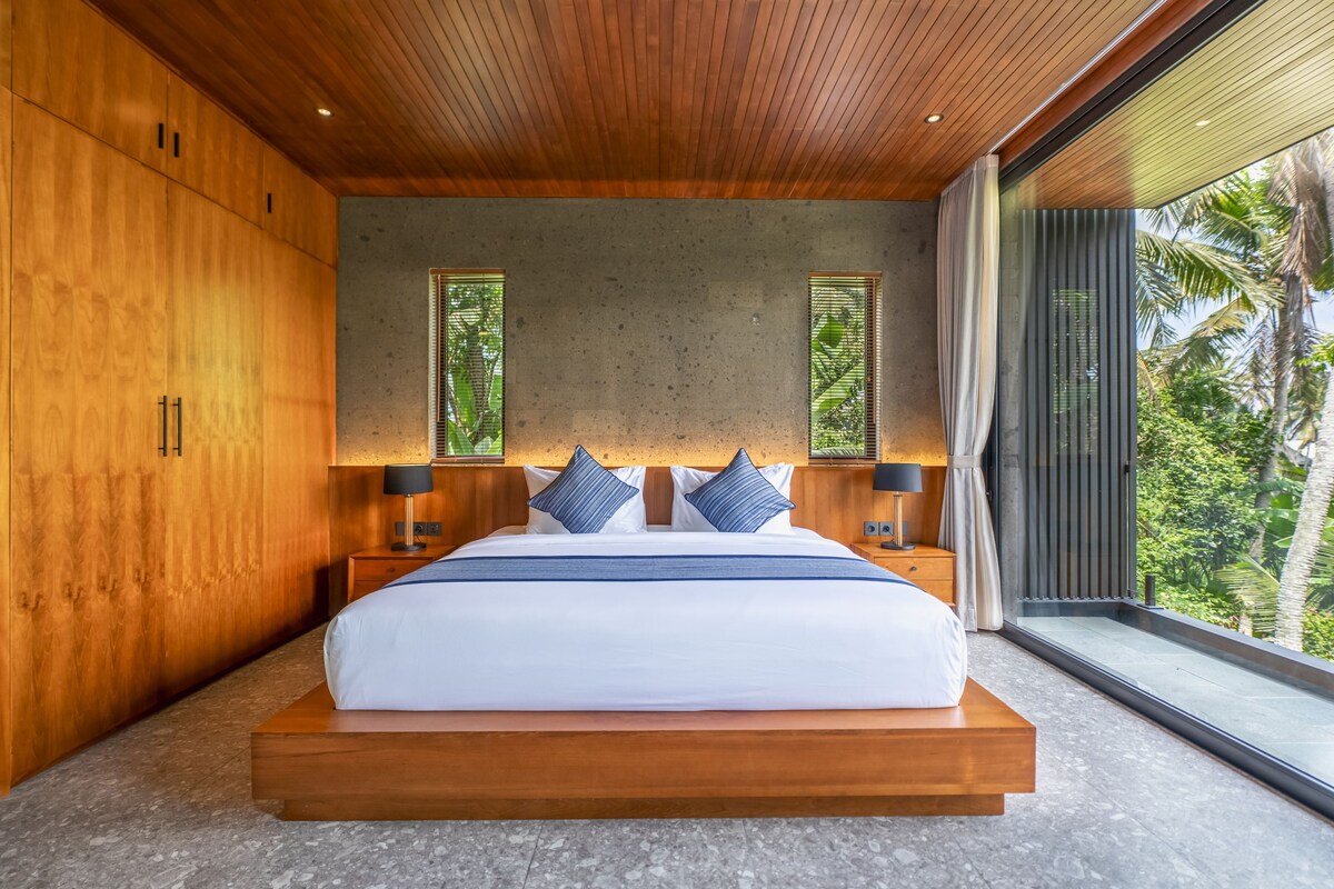 A modern bedroom with a large bed, wooden walls, and a view of greenery.