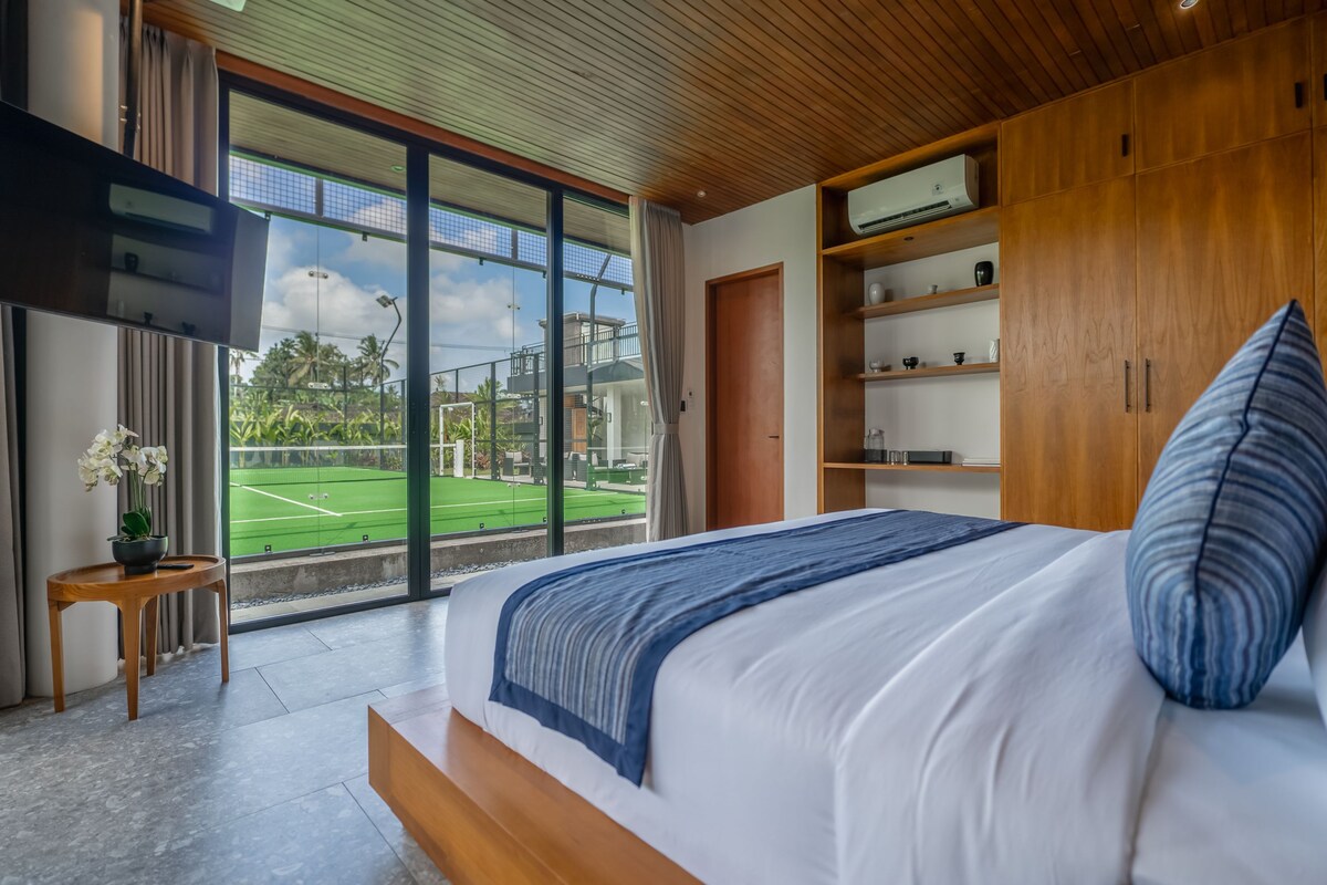 A bedroom with a large bed, wooden headboard, and a view of a tennis court through floor-to-ceiling windows.