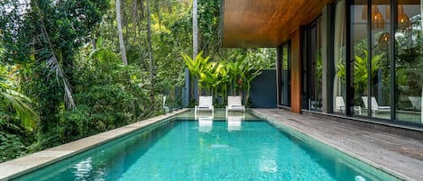 Una piscina cristalina con sillones de descanso, rodeada de exuberante vegetación y un edificio moderno con grandes ventanales.