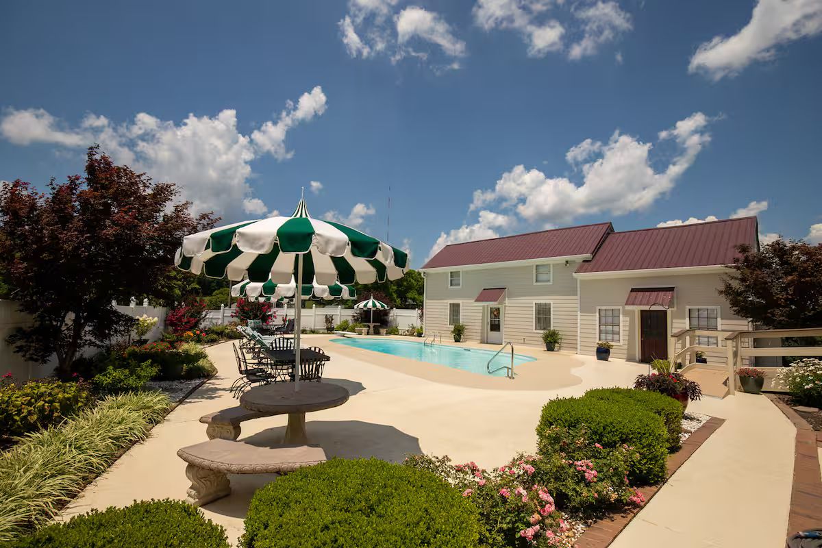 A pool area with a lounge chair and umbrella, surrounded by well-maintained landscaping and a building with a red roof.