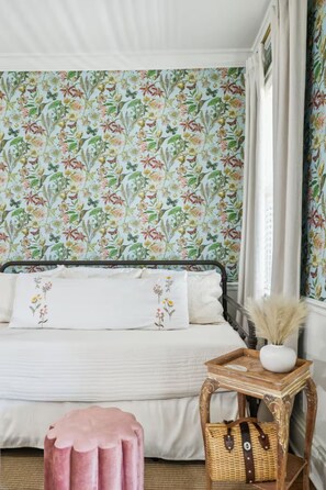 Quarto com papel de parede floral, cama com roupa de cama branca, pequena mesa de cabeceira de madeira com um vaso e um banquinho rosa.