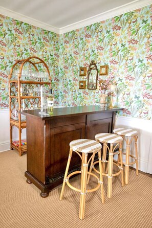 A bar area with a wooden bar counter, three wicker stools, a glass display case with glasses, and a floral wallpaper background. - The Pearl Retreat (Nashville)