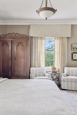 A bedroom with a large bed, two armchairs, a wooden wardrobe, a window with curtains, and a hanging light fixture. - The Pearl Retreat (Nashville)