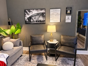 A hotel room with a sofa, two chairs, a lamp, a framed picture of a stadium, and informational posters on the wall. - Destino Stadium Suite 428 (Arlington)