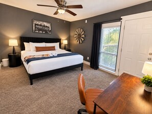 A hotel room with a bed, a desk, a chair, a ceiling fan, a window with blinds, and a decorative wall piece. - Destino Stadium Suite 428 (Arlington)