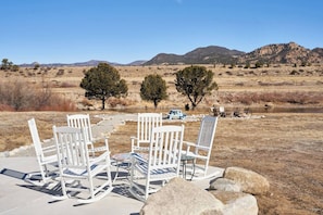 A patio with white rocking chairs and a table, overlooking a dry landscape with sparse trees and distant mountains.