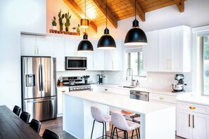 Una cucina moderna con mobili bianchi, elettrodomestici in acciaio inox, un'isola centrale con sgabelli da bar, illuminazione a sospensione e soffitto in legno.