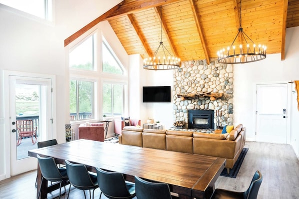 A spacious living area with a large wooden dining table, black upholstered chairs, a stone fireplace, and a television mounted on the wall.