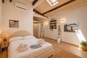 Quarto com cama, mesa de cabeceira com abajur, ar-condicionado fixado na parede, escada, prateleira com objetos decorativos, espelho e quadro emoldurado.