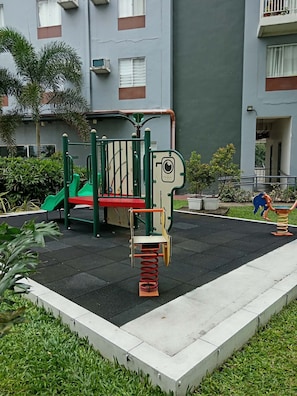 A playground with a seesaw, slide, and climbing structure, surrounded by a paved area and greenery.