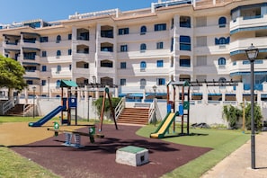 A playground with slides and swings, surrounded by a building with multiple balconies and a clear sky. - Manilva Beachfront Promenade by Libehomes (San Luis De Sabinillas)