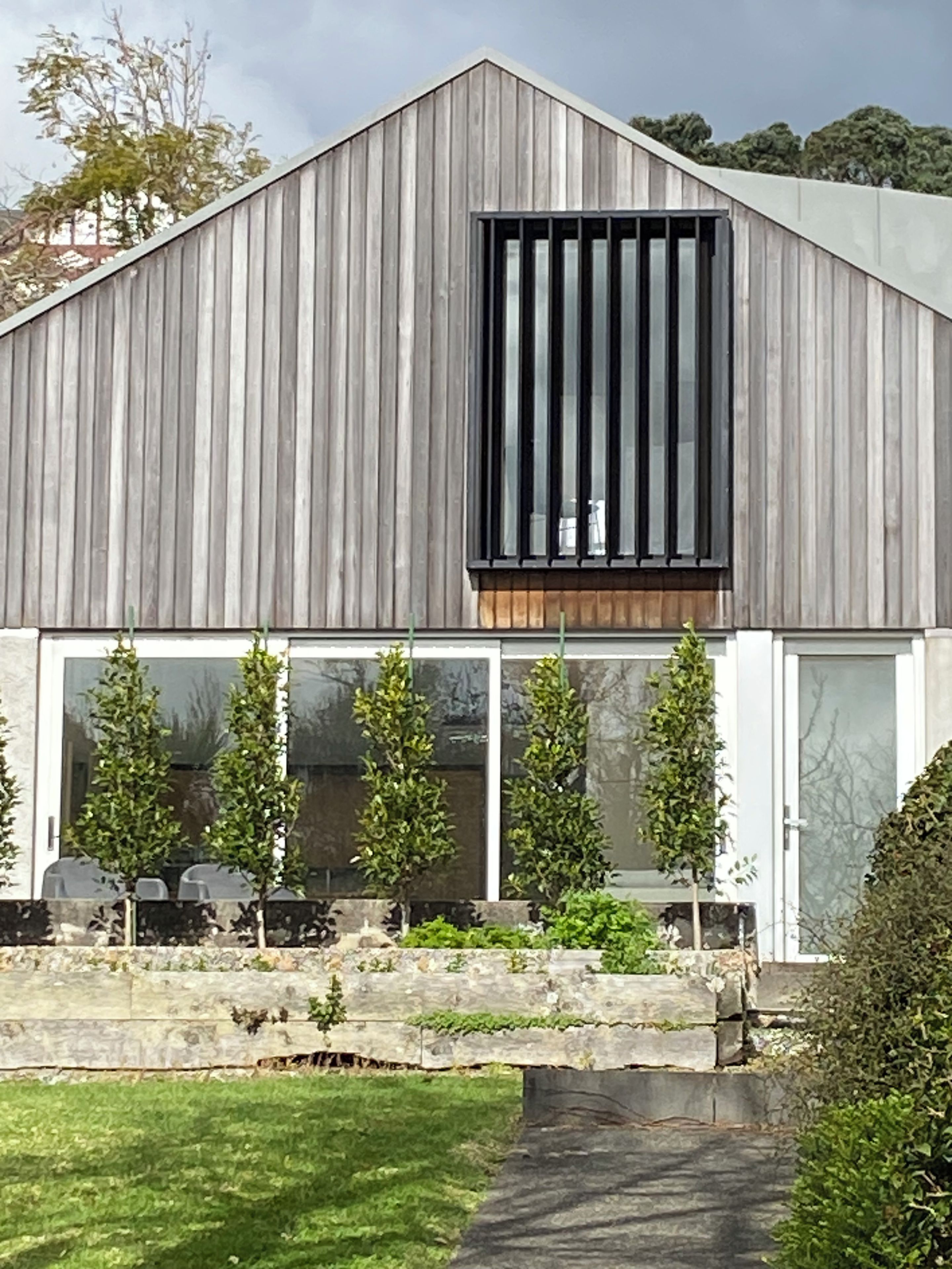 A modern building with a sloped roof, large windows, and a stone retaining wall with greenery.