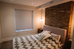 Une chambre à coucher comprenant un lit, une table de chevet, une fenêtre avec des stores et un mur décoratif en briques.