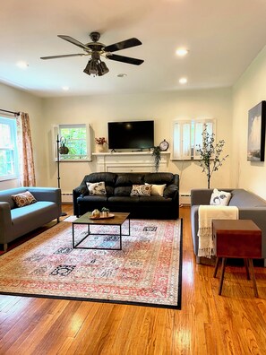 A living room with a black sofa, a grey sofa, a coffee table, a ceiling fan, and a television on the wall. - Peaceful Retreat with Massive Private Deck – Free Parking & Easy Highway Access (Vestal)