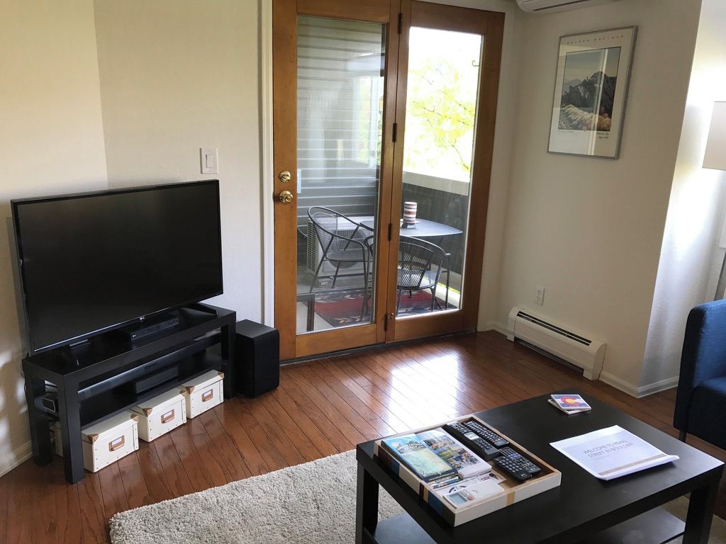 Un salon avec un téléviseur à écran plat posé sur un meuble, une table basse avec des livres et des télécommandes, une porte-fenêtre coulissante donnant sur un balcon aménagé avec du mobilier d’extérieur, et un tableau encadré accroché au mur.