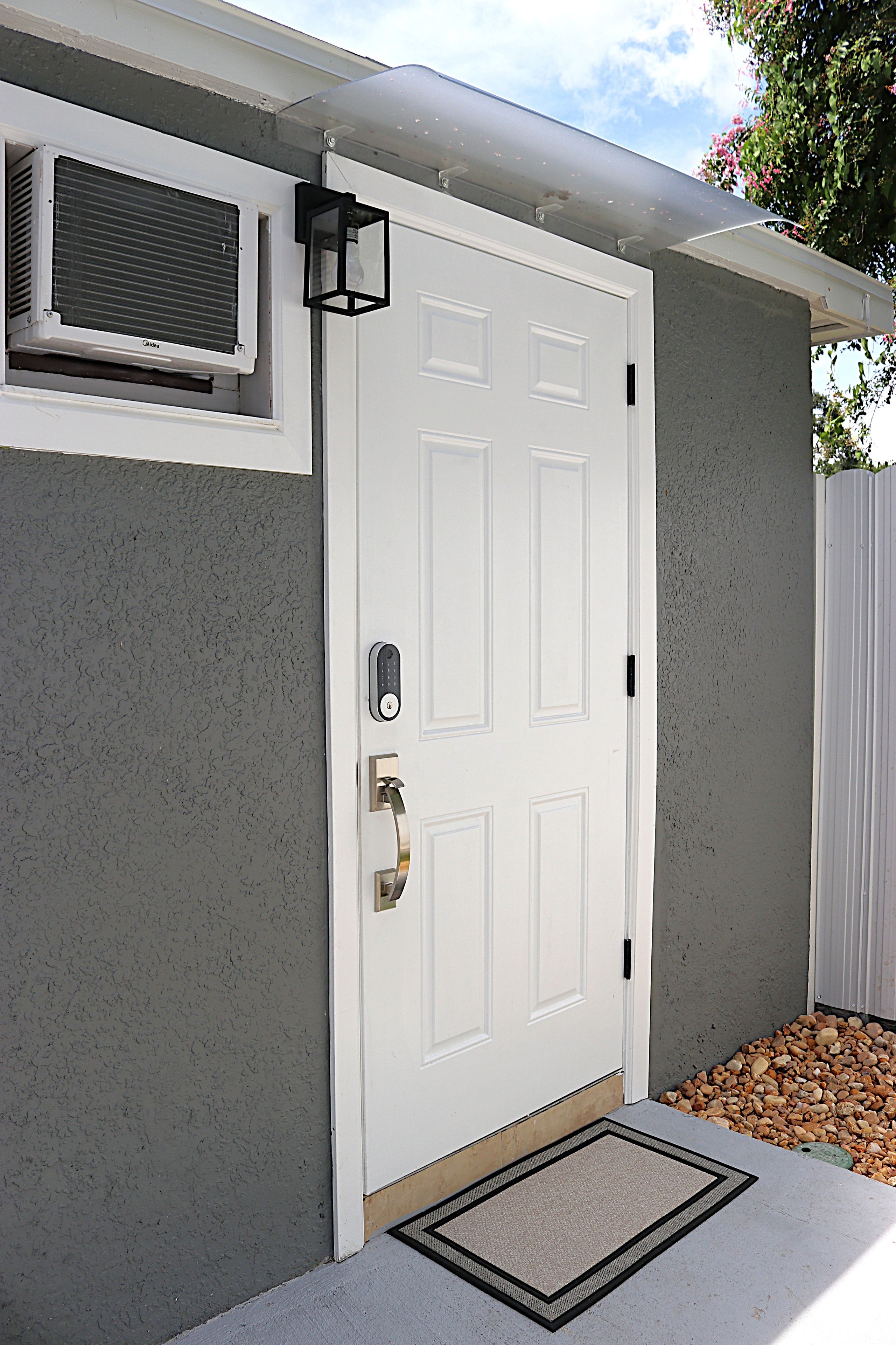 A white door with a keypad lock, a small window with a vent, and a doormat on a concrete surface.