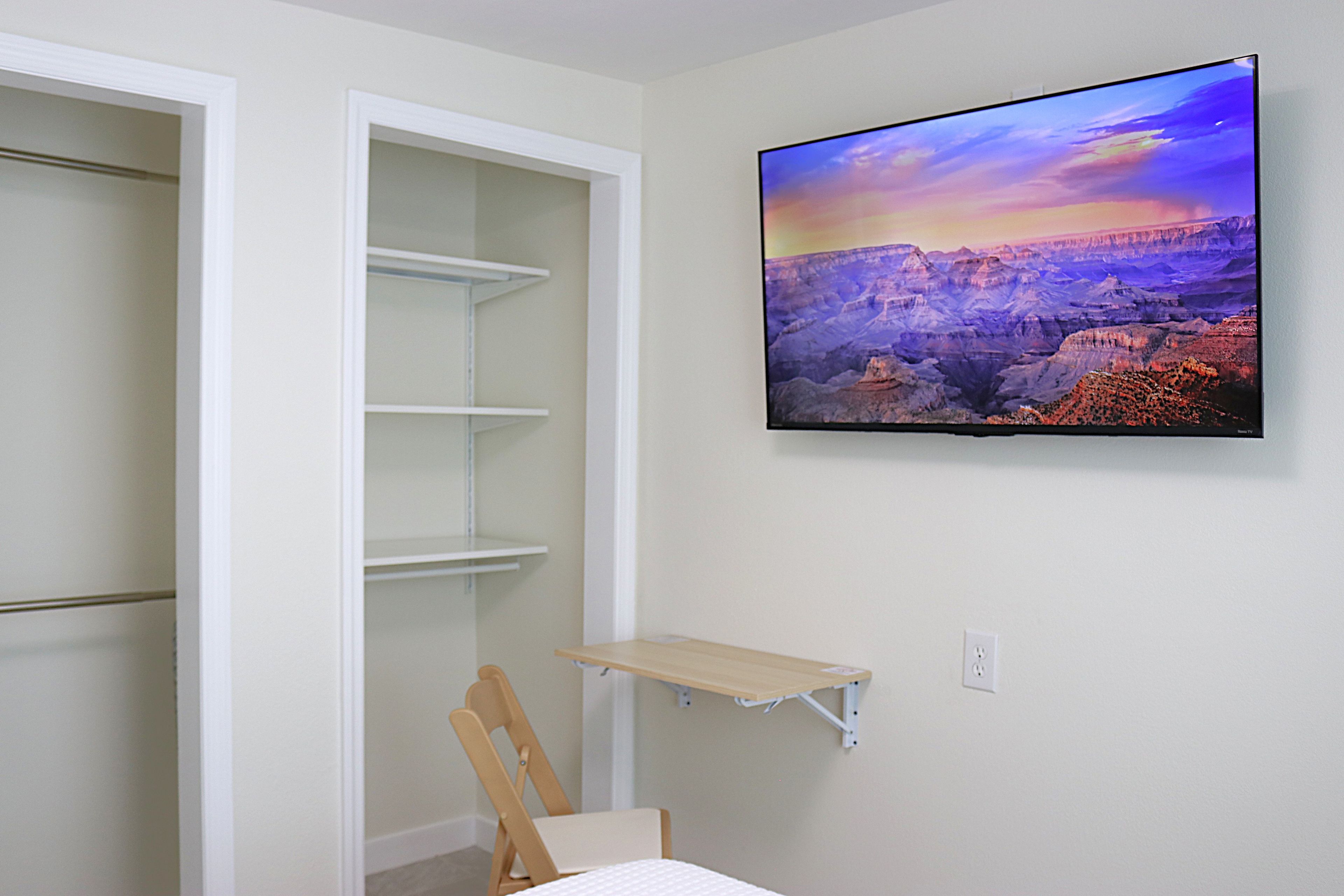 A room with a wall-mounted TV displaying a scenic landscape, a wooden chair, and a small table.