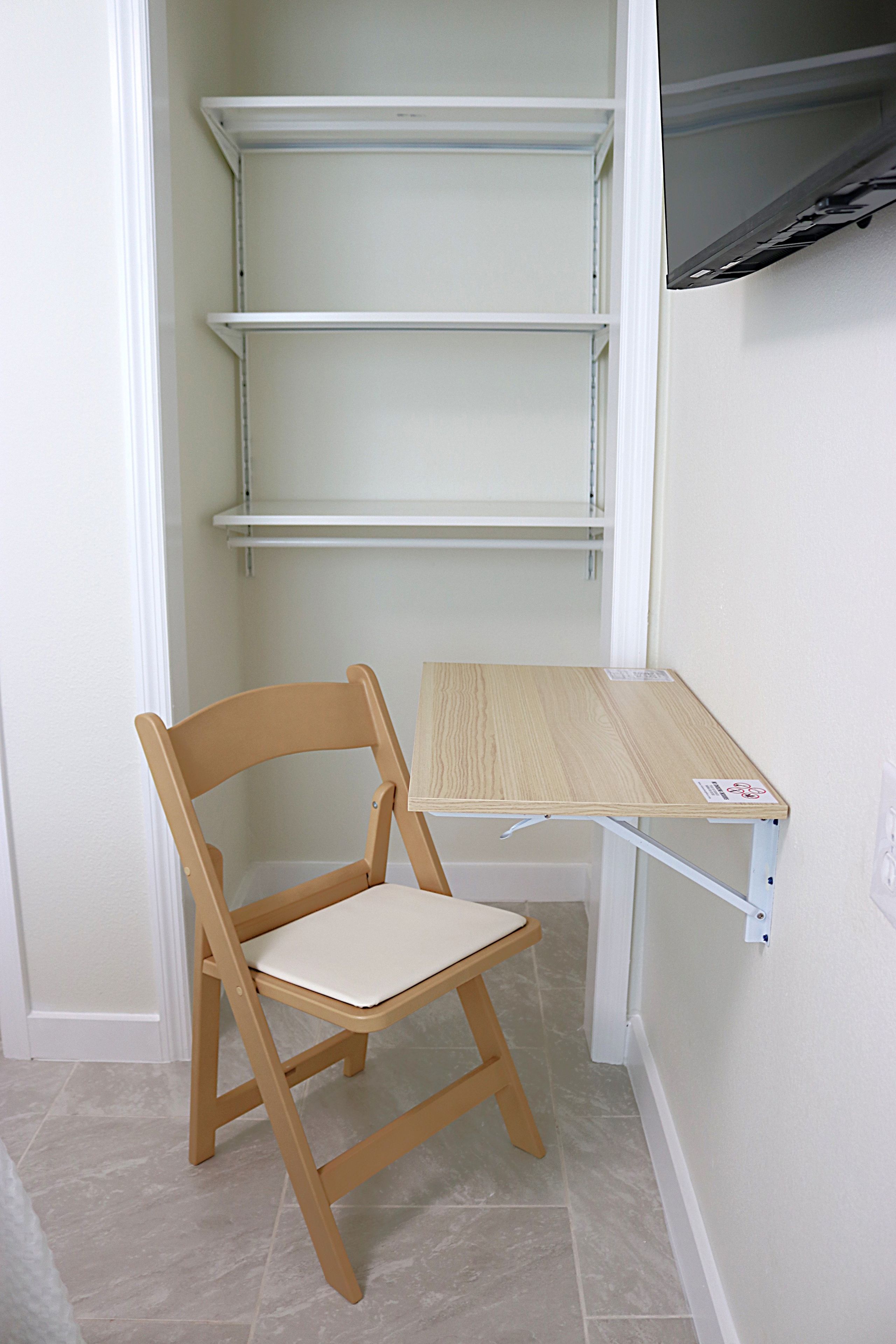 A wooden chair with a cushion and a small wooden table with a label, positioned in front of a white wall with a shelf unit above.