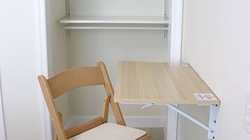 A wooden chair with a cushion and a small wooden table with a label, positioned in front of a white wall with a shelf unit above.