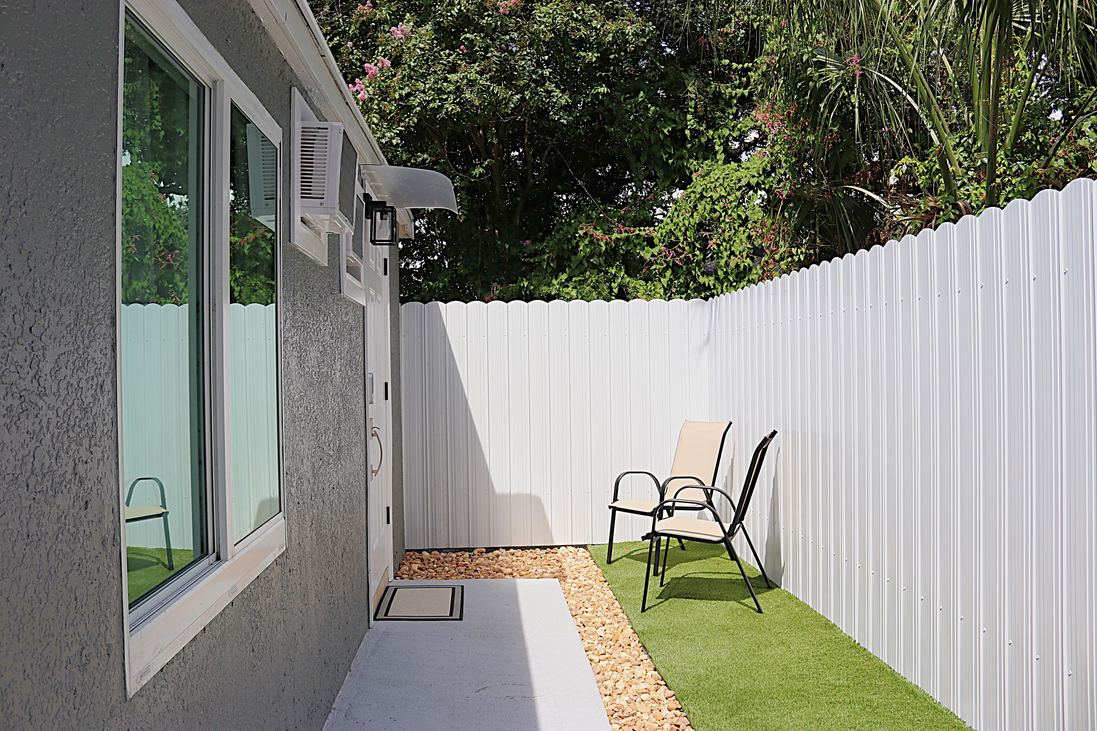 A patio with a white fence, a small seating area with chairs, and a window with an air conditioning unit.