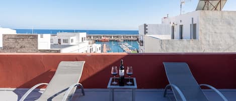 A rooftop terrace with two lounge chairs, a table with a bottle of wine and glasses, and a view of a marina with boats and the sea.