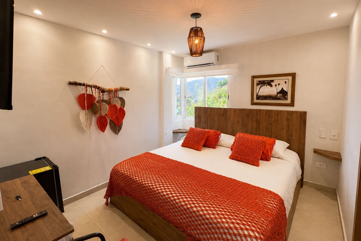 A bedroom with a bed, a wooden headboard, a wall-mounted air conditioner, a window with a view, and a wall decoration.