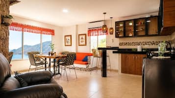 Ein modernes Wohnzimmer mit Sofa, Essbereich, Küche und großen Fenstern mit Blick auf die Berge.