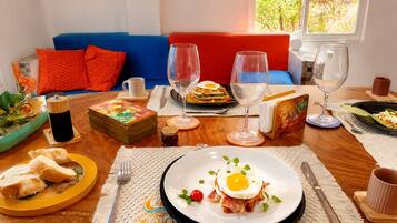 A dining table set with plates of food, glasses, and cutlery, a blue sofa with red cushions, and a window with a view of greenery.