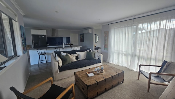 A living room with a sofa, coffee table, and chairs. A kitchen is visible in the background. - Relax in Beautiful Papamoa – Stylish, Comfortable Home Close to the Beach (Tauranga)