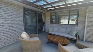 An outdoor patio with a glass door leading to a dining area, a sofa, and wicker chairs. - Relax in Beautiful Papamoa – Stylish, Comfortable Home Close to the Beach (Tauranga)