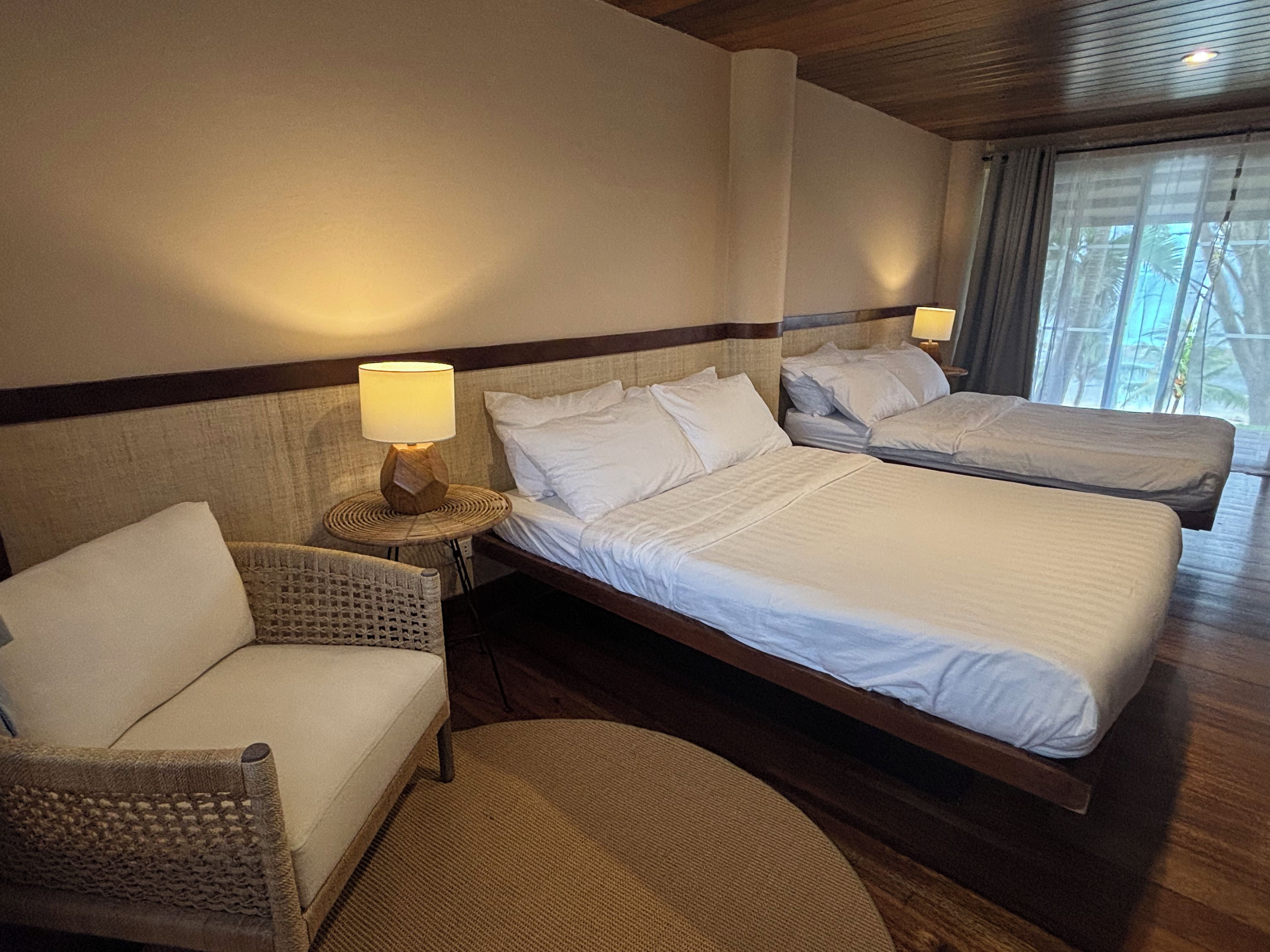 Habitación de hotel con dos camas, una silla, una lámpara y vistas a palmeras a través de la ventana.
