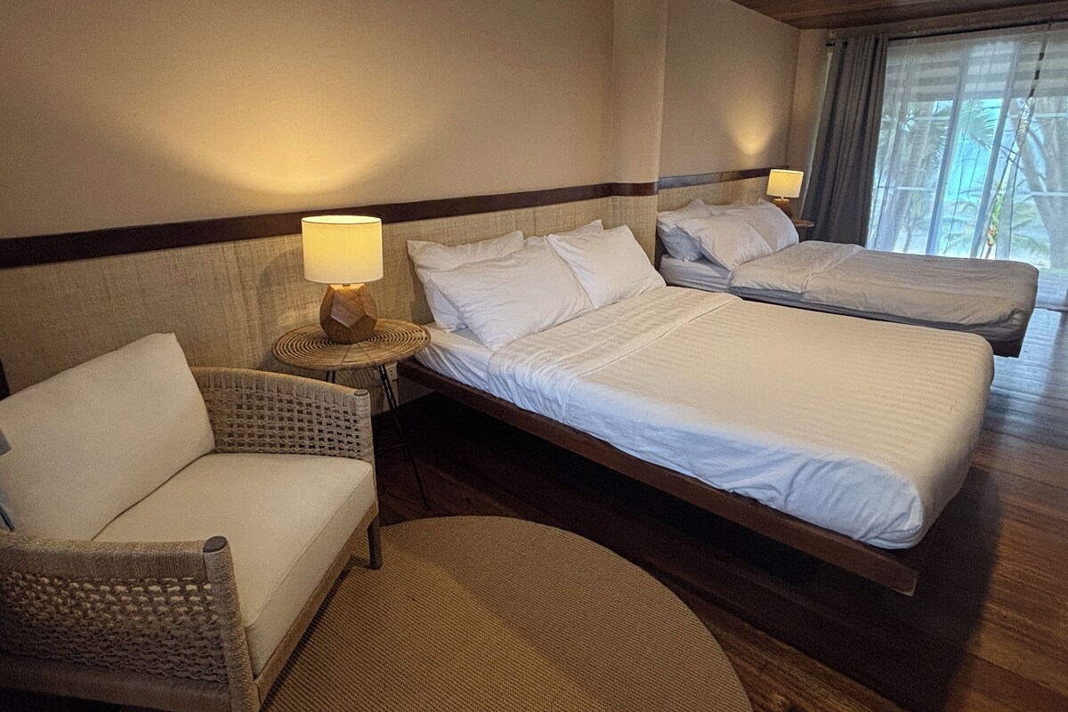 Habitación de hotel con dos camas, una silla, una lámpara y vistas a palmeras a través de la ventana.